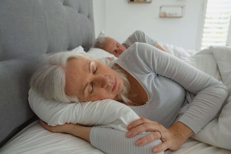 3 Health Benefits for Using Your AC While Sleeping. Photo of a senior couple sleeping soundly in their bed.