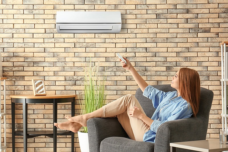 Hot and Cold Spots? Get Even Heating and Cooling in Your Home. Photo of a woman sitting on a chair in her home, adjusting the AC with a remote.
